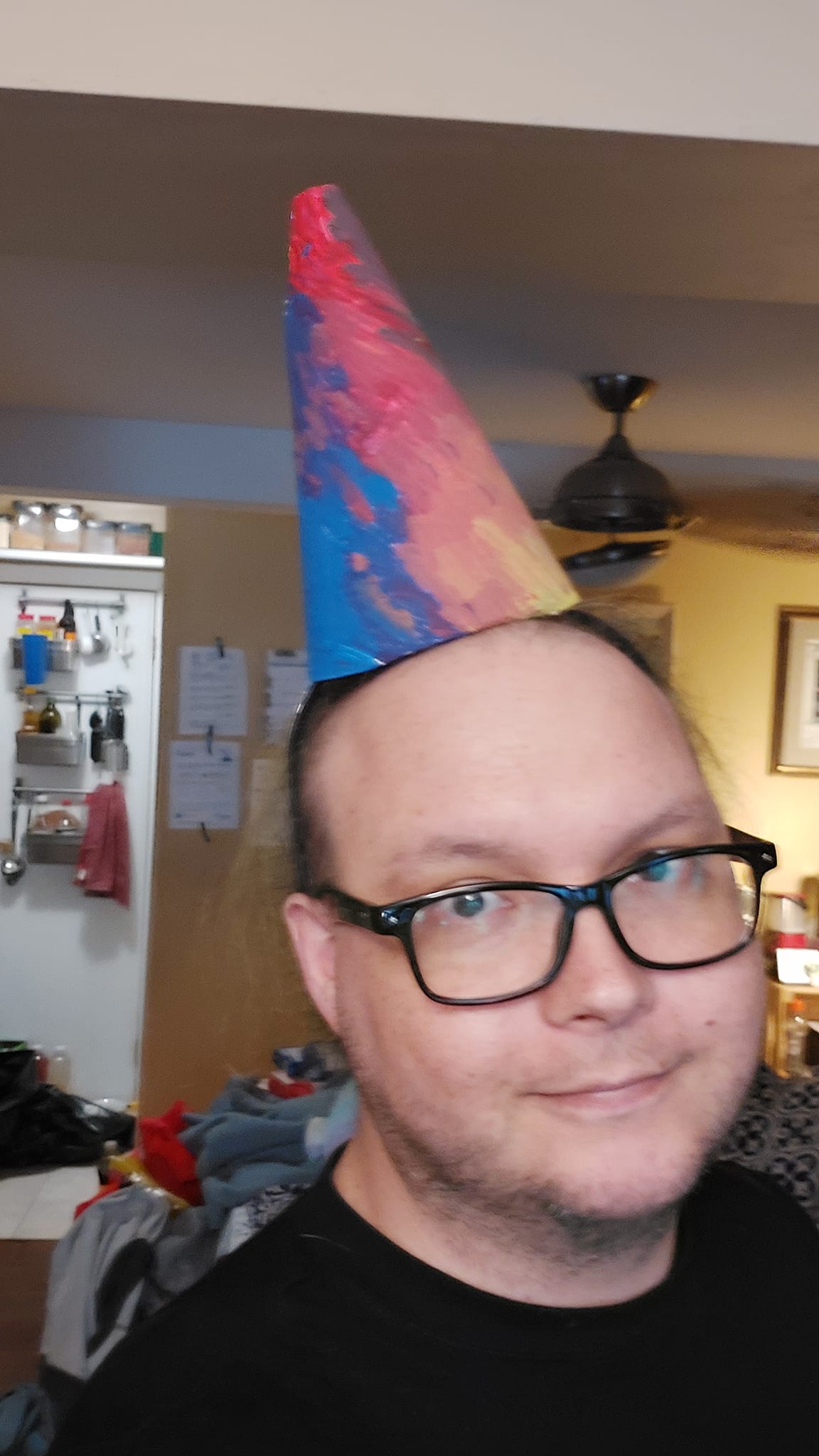 A man sits in a computer chair, wearing a hand painted party hat and a black shirt